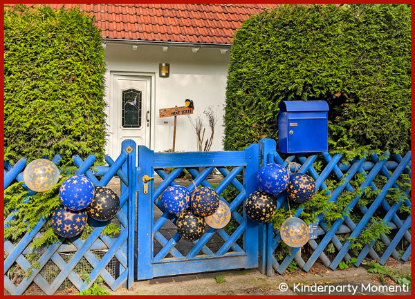 an blauen Holzzaun hängen blaue Luftballons mit goldenen Sternen, im Hintergrund ein Haus