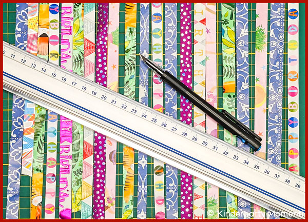 Streifen von Geschenkpapier auf einer Bastelmatte, darauf eine Lineal und ein Schenidemesser