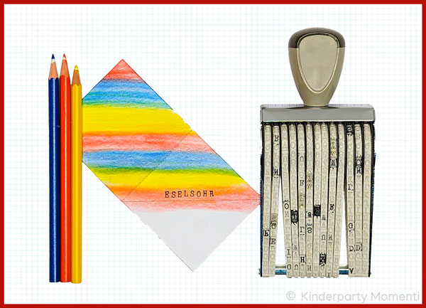 drei Buntstifte, ein Buchstabenstempel und ein in regenbogenfarben bemaltes Papier mit dem Aufdruck Eselohr