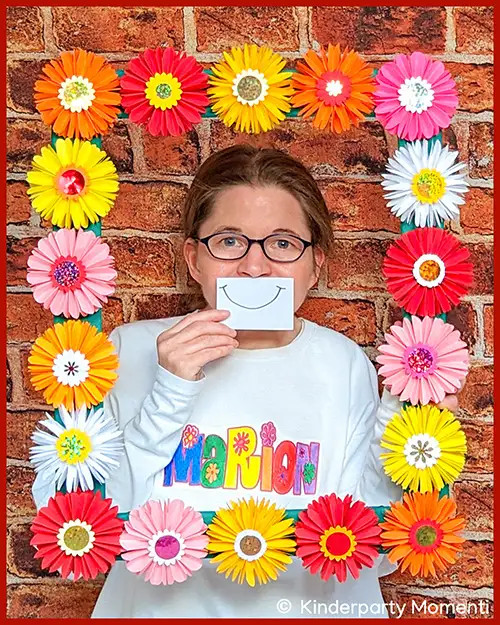 Marion Seike - Freizeitpädagogin und Autorin der Website Marion von Kinderparty Momenti hält ein Schild mit einem lächelndem Mund vor ihr Gesicht und trägt ein weißes Sweatshirt mit buntem Schriftzug 'Marion', umrahmt von einem Rahmen aus großen Papierblumen in verschiedenen Farben im Hintergrund eine Ziegelsteinwand